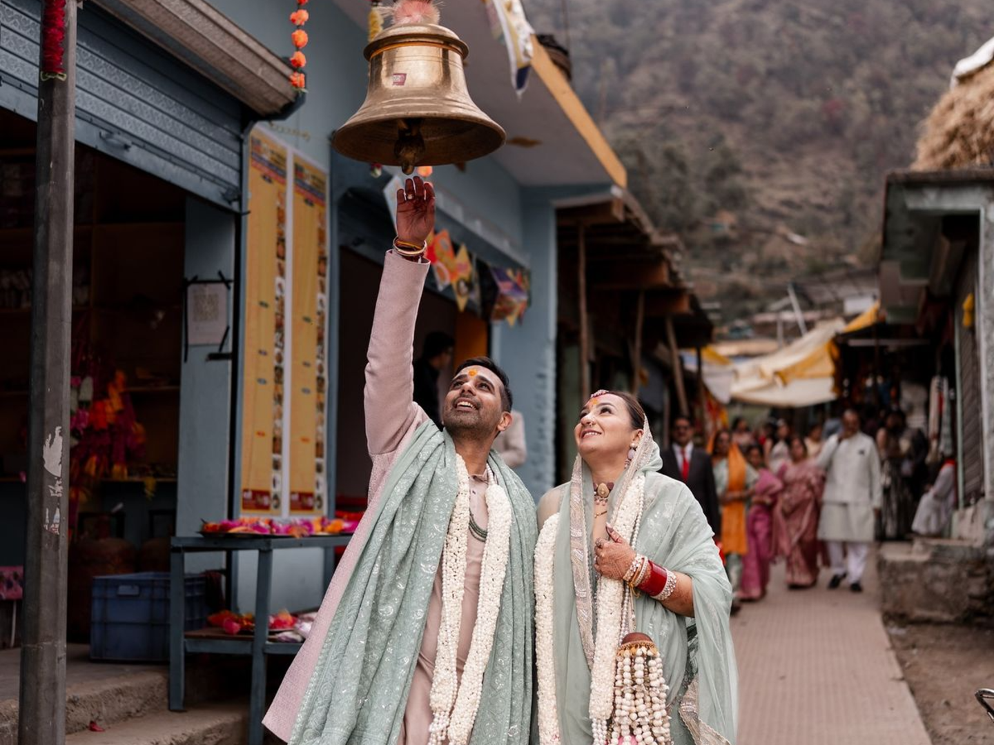 A candid, joyful photograph of the Indian bride and groom ringing a large brass bell in a street market setting near a temple in the Uttarakhand mountains. The groom, wearing a pale pink and green outfit, reaches up, while the bride, in a light mint green sari and heavily adorned with floral garlands (varmala), looks up at him smiling. This captures a unique, intimate moment during their destination wedding, similar to the simple elegance seen in ceremonies like Samantha Ruth Prabhu's.